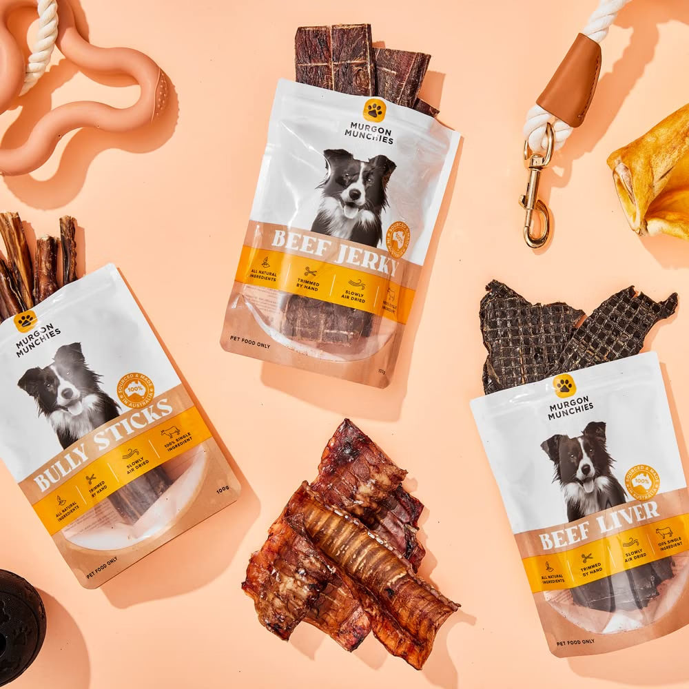 Three bags of Mission Dogs & Humans dog treats arranged on a peach background with the treats themselves scattered around. The bags are labeled Billy Sticks, Beef Jerky, and Beef Liver, each showing a black-and-white dog portrait and orange-and-white branding.