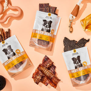 Three bags of Mission Dogs & Humans dog treats arranged on a peach background with the treats themselves scattered around. The bags are labeled Billy Sticks, Beef Jerky, and Beef Liver, each showing a black-and-white dog portrait and orange-and-white branding.