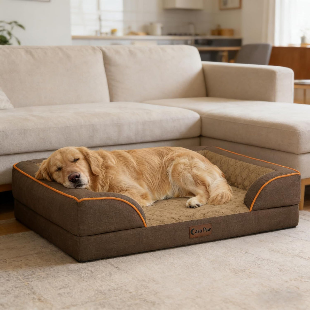A golden retriever dog rests on a brown raised pet bed with orange piping in a living room setting with a beige sectional sofa in the background. The brand name 'Cula Paw' is visible on a small leather patch on the front of the bed at Pet Servo