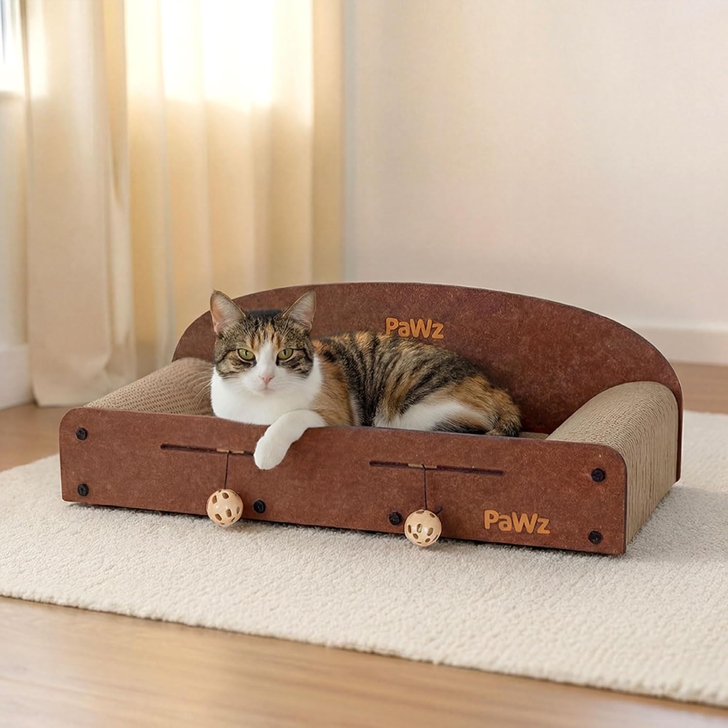 A calico cat lies inside a brown and beige PawWz cat bed with dangling toy balls, placed on a cream rug in a softly lit room at Pet Servo