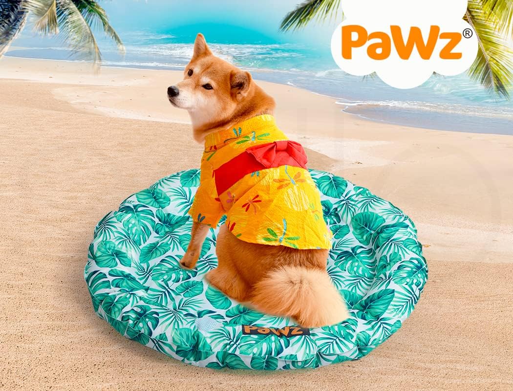 A Shiba Inu dog wearing a yellow Hawaiian shirt sits on a round green and white palm-leaf patterned dog bed on a sandy beach with the ocean and palm trees in the background. The PaWz brand logo is visible in the top right corner at Pet Servo