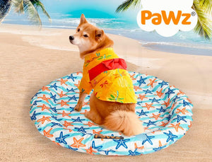 A small dog sits on a round beach mat with a starfish pattern on a sandy beach, wearing a yellow and red Hawaiian shirt. The PaWz brand logo is visible in the top right corner at Pet Servo