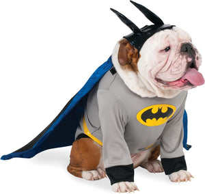 An English Bulldog wearing a Batman costume, featuring a gray long-sleeve shirt with a yellow Batman logo, black cuffs, a blue cape, and a black bat-shaped headband, photographed against a solid white background at Pet Servo