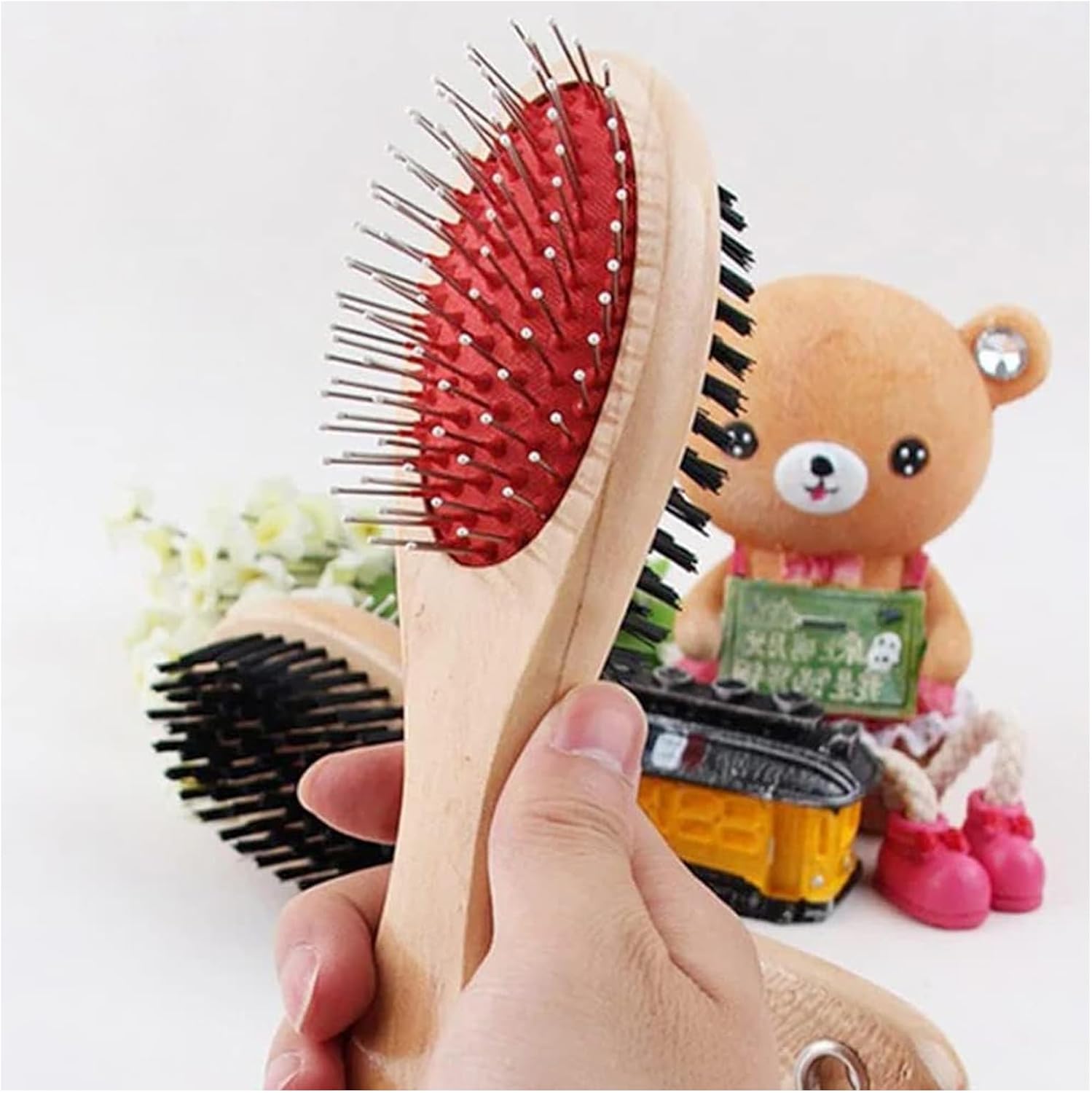 Hand holding a wooden hairbrush with red bristle head, teddy bear toy in the background at Pet Servo