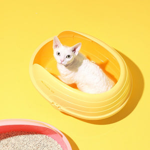 A white cat sits inside a bright yellow oval plastic litter box on a solid yellow background, with a partial pink litter box and litter visible in the lower left corner at Pet Servo