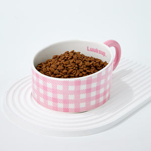 A pink and white checkered pet bowl filled with brown kibble, placed on a white ridged tray against a clean white background. The inside of the bowl shows the brand name 'Luuksup' at Pet Servo