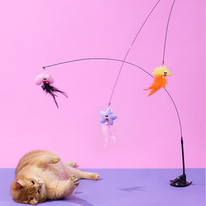 A ginger cat lies on its back on a purple surface, looking at a black cat toy with three dangling colorful plush toys against a pink and purple studio background at Pet Servo