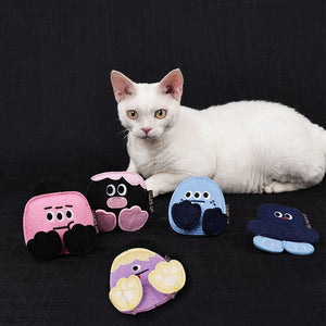 A white cat lies on a black fabric background behind several small plush character pouches in pink, black, blue, purple, and navy. The pouches have cartoon faces and are arranged around the cat at Pet Servo