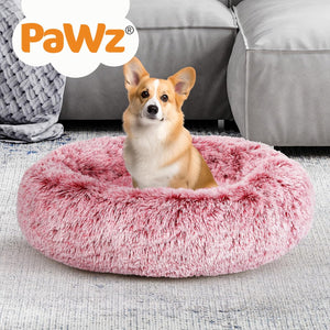 A tan and white Corgi sitting in a fluffy pink round pet bed on a light gray rug, with a gray sofa in the background and a white cloud-shaped PaWz logo in the top left corner at Pet Servo