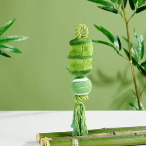 A green dog toy made from a tennis ball and plush fabric, hanging from a rope loop, displayed on bamboo poles against a green background with blurred plants at Pet Servo