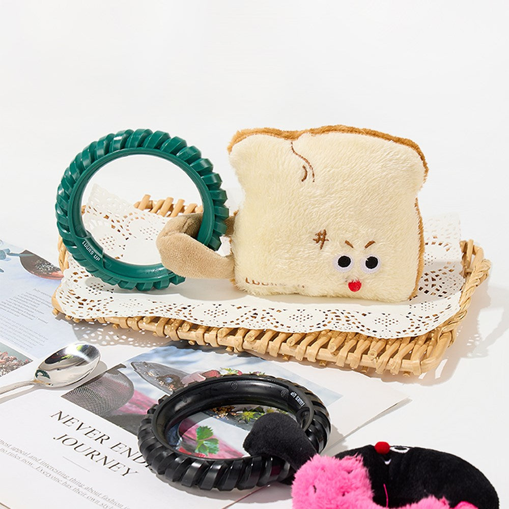 A plush bread-torte character pouch sits on a woven tray with a lace pad, alongside a green circular ring, a black ring, a spoon, and a magazine on a white background at Pet Servo