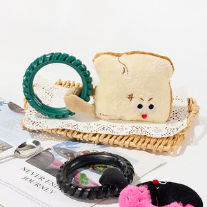 A plush bread-torte character pouch sits on a woven tray with a lace pad, alongside a green circular ring, a black ring, a spoon, and a magazine on a white background at Pet Servo