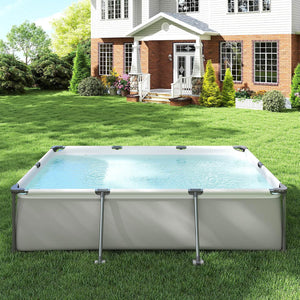 A rectangular above-ground swimming pool with a light gray frame and clear blue water, set on a green lawn in front of a white and brick house.