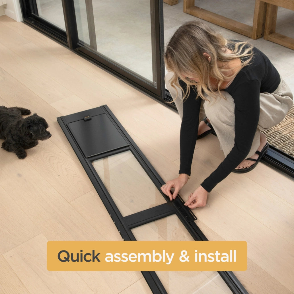 A woman kneels on a light wood floor assembling a black-framed sliding glass door panel next to a small black dog. A yellow text banner at the bottom reads 'Quick assembly & install' at Pet Servo