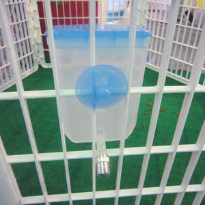 A clear plastic pet waterer with a blue lid and blue suction cup attachment, mounted on a white plastic playpen over green artificial turf at Pet Servo