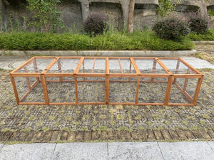 A long wooden frame structure with clear panels, photographed outdoors on a stone-paved surface with shrubs and a stone wall in the background at Pet Servo