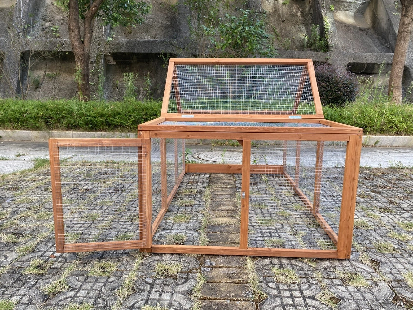 A wooden chicken coop with wire mesh panels, featuring an open side door and a hinged top lid, set outdoors on a paved stone surface with greenery and a rock wall in the background at Pet Servo