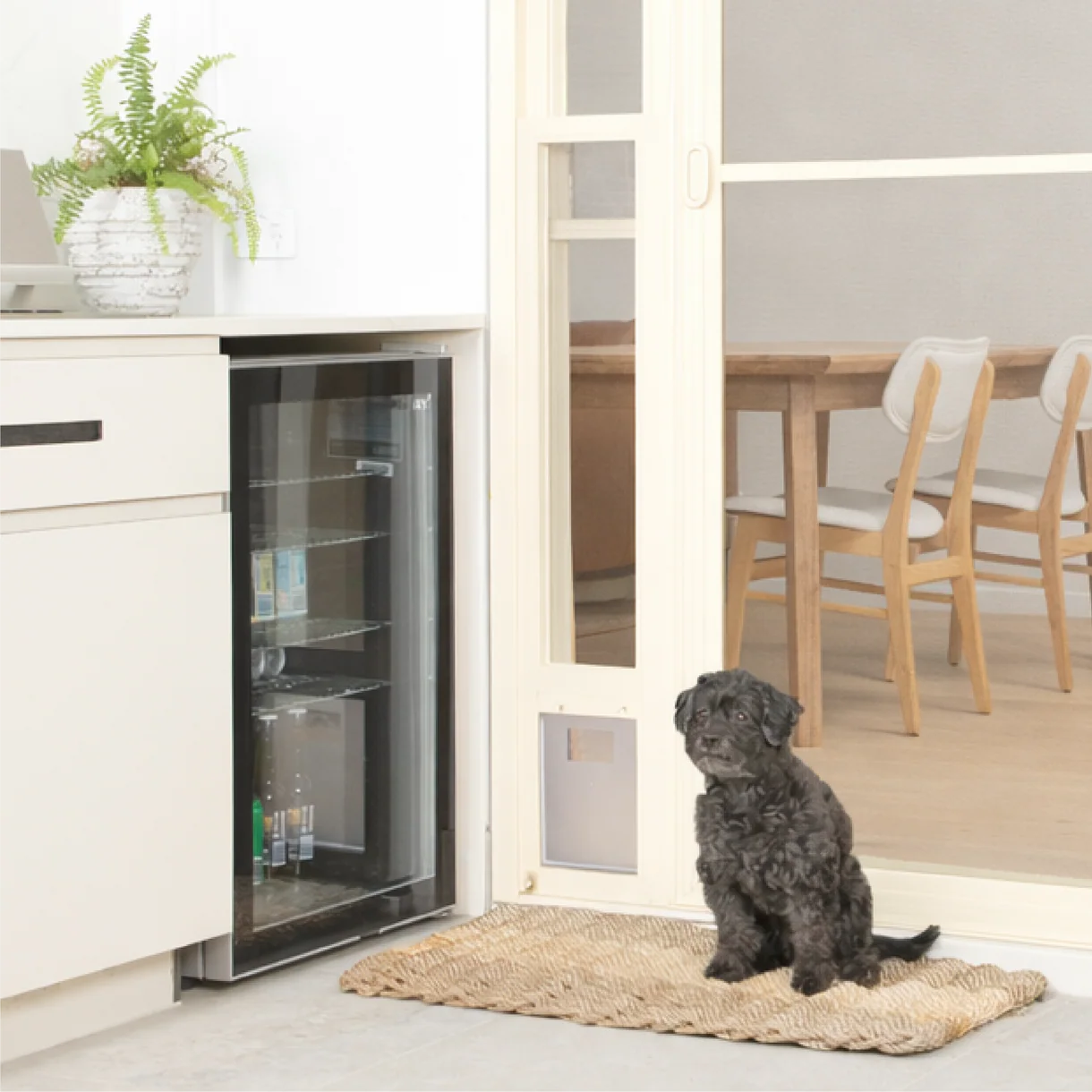 Dog sitting on a mat in front of a glass door pet door in a kitchen.