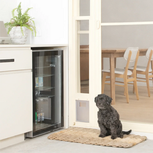 Dog sitting on a mat in front of a glass door pet door in a kitchen.