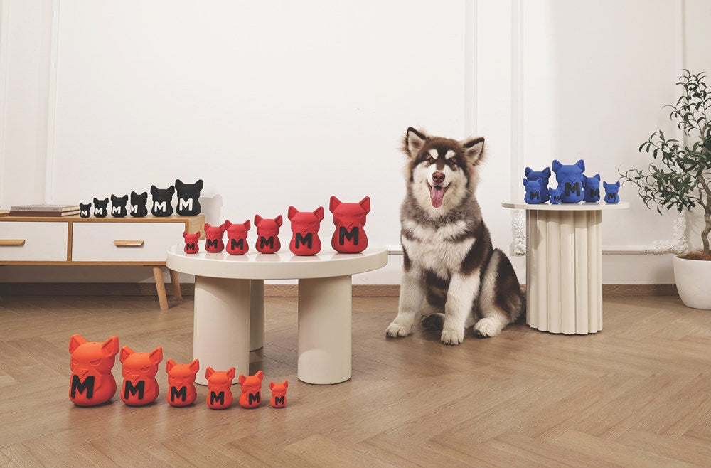 A fluffy black, white, and gray dog sits on a light wood floor in a bright room, surrounded by multiple red and blue plush cat-shaped toys with embroidered letters 'M' and 'C' on them. The scene includes low white tables, a cabinet, and a potted plant at Pet Servo