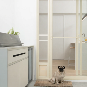 Pug sitting on a mat in front of a glass door with a view into a modern kitchen.