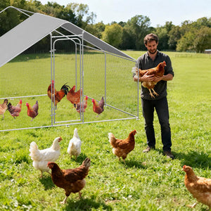 Man holding a chicken next to a metal chicken coop in a grassy field with chickens around.