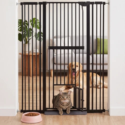 A black metal pet gate with a low-profile door and a small ramp, shown installed in a doorway in a home setting. A brown tabby cat is sitting on the ramp in the foreground, and a golden retriever dog is lying on the floor behind the gate at Pet Servo